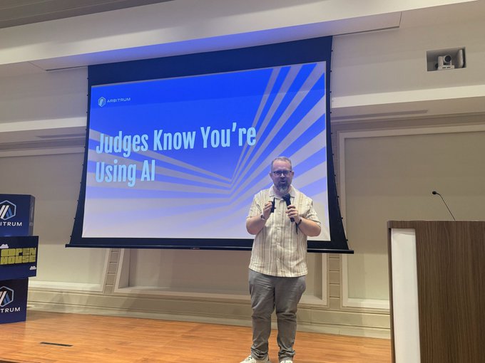 A man giving a presentation on stage holding a microphone, with a projection screen behind him displaying the title 'Judges Know You're Using AI' at an Arbitrum event.
