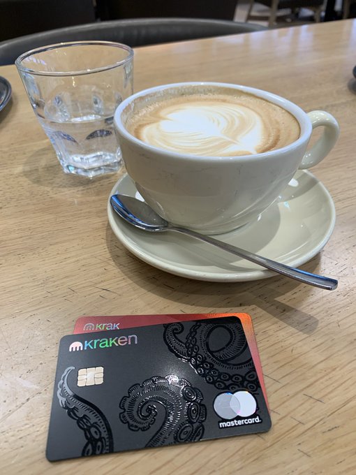 A flat lay on a wooden table showing a cappuccino in a white cup, a glass of water, and a black 'kraken' Mastercard with octopus design.