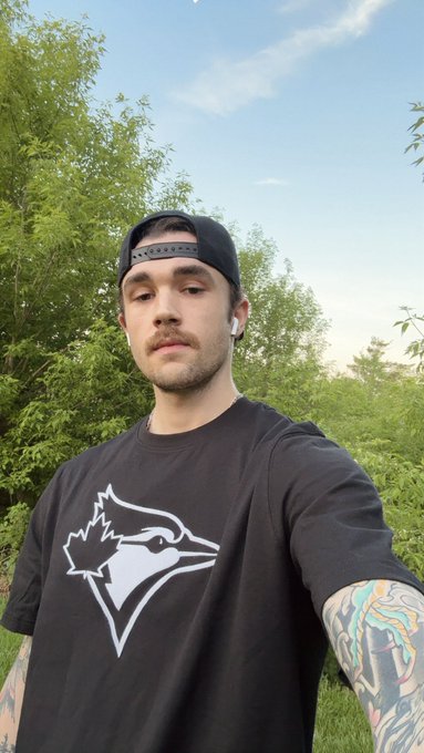 A young man with a beard, backwards black cap, and earbuds takes a selfie outdoors wearing a Toronto Blue Jays t-shirt, with a tattooed arm visible, introducing himself as the person behind the account.