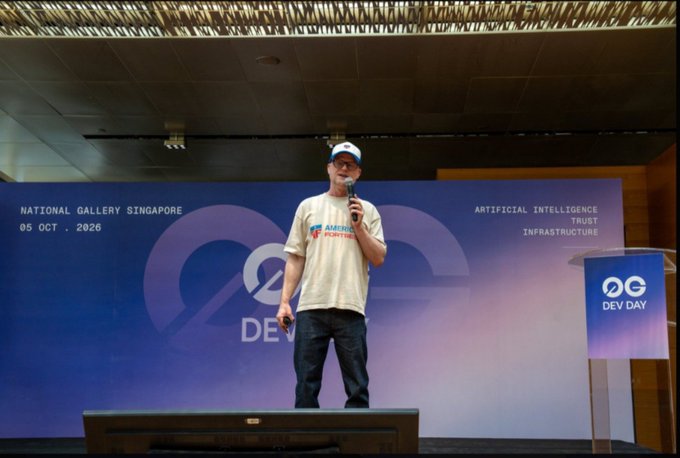 A man wearing a cap and "AMERICAN FORTRESS" t-shirt speaks at a podium during the "DEV DAY" event at the National Gallery Singapore on 05 OCT 2026, with banners highlighting "Artificial Intelligence, Trust, Infrastructure." The image ties to the accompanying post by showcasing a presenter from American Fort(io) discussing their approach to agent payments, which balances verification with wallet privacy—addressing the very tradeoff between proof of payment and exposure noted in the commentary.