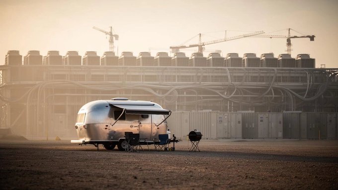 A silver Airstream-style travel trailer is parked on dirt with camping chairs set up beside it, set against an industrial backdrop with construction cranes and hazy lighting.
