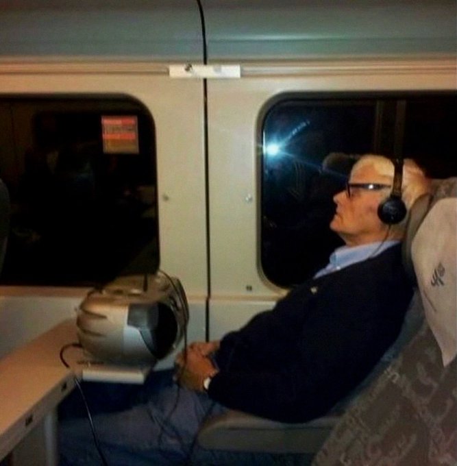 An older man with glasses and headphones sits on a train at night with a portable fan pointed at him.
