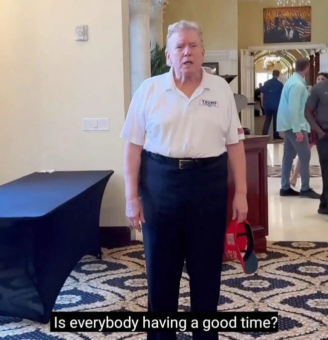 Donald Trump wearing a white polo shirt with a "TRUMP" logo stands in a hotel-like setting asking "Is everybody having a good time?"