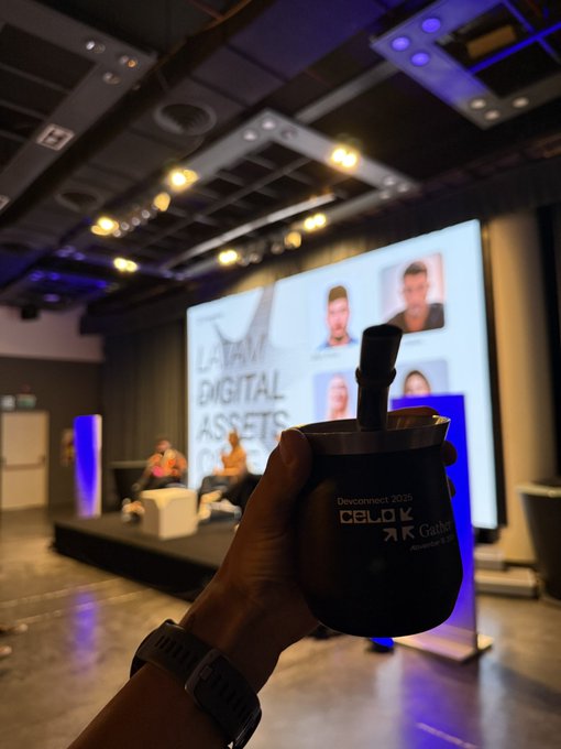 A hand holding a traditional yerba mate gourd at a 'LATAM Digital Assets' conference, with a presentation screen showing panelists in the background.