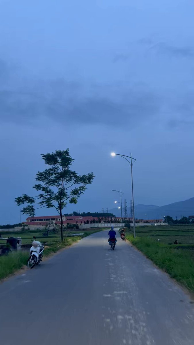 The image shows a road with two motorcycles parked next to each other, one of which has its headlights on. There are three people riding their bikes down the street, with one person closer to the left side of the scene and another person further back in the right lane. The background features a cloudy sky and trees lining the road, creating an atmosphere of dusk or dawn.