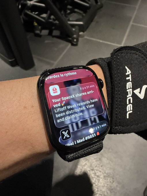 The image shows a person wearing a black wristwatch that displays an email message from a company named "Atecel". The watch has a black strap, which contrasts with its silver face. The watch's screen shows a message about rewards and discounts for using a specific app or service. The person is also wearing a black glove on their hand.