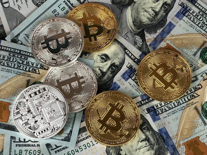 The image features a collection of Bitcoins scattered across a table with various denominations of US dollar bills. The Bitcoins are displayed prominently, while the dollar bills are positioned underneath them. This arrangement suggests that someone might be researching or discussing Bitcoin investments, given their close proximity on the table and the presence of money in the background.