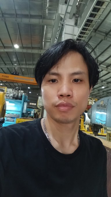 A man with dark hair wearing a black shirt takes a selfie inside what appears to be a factory or industrial facility with a high ceiling and overhead crane.