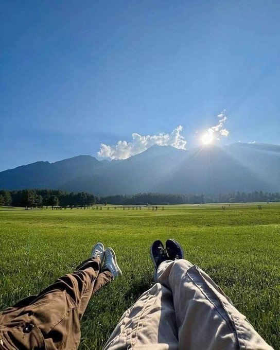 The image shows two people sitting on a grassy field, with their legs crossed, facing each other. They are wearing sneakers and pants, suggesting they might be taking a break from an outdoor activity or enjoying some leisure time together. The sun is shining brightly in the sky, casting shadows and highlighting the beauty of nature around them.