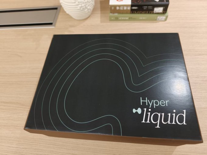 A black gift box with teal wave-like designs and the 'Hyperliquid' logo sits on a wooden desk near some books and a white textured cup.