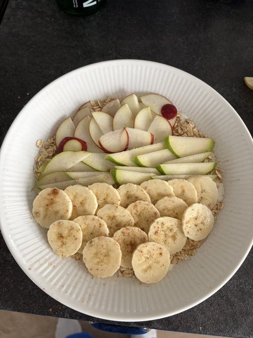 A white ridged bowl on a dark countertop contains a healthy breakfast of oatmeal topped with neatly arranged slices of apple, green pear, and banana, dusted with cinnamon.