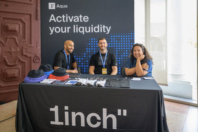 Three people sitting behind a black tablecloth that reads '1inch', with a banner behind them displaying 'Aqua' and 'Activate your liquidity'.