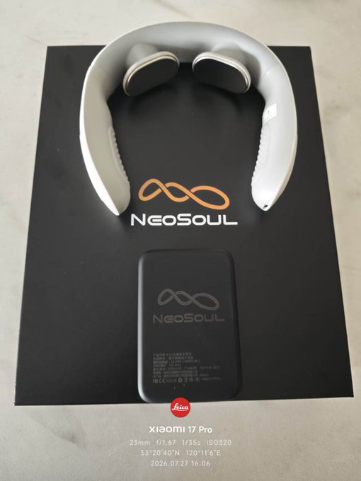 A flat lay photograph of a white NeoSoul neck massager and its black charging case on a branded box, watermarked as taken with a Xiaomi 17 Pro on July 27, 2026.