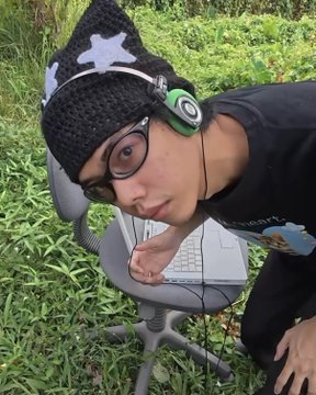 A young man wearing glasses, a knitted black beanie with a white star, and green headphones poses outdoors next to a laptop on a chair.