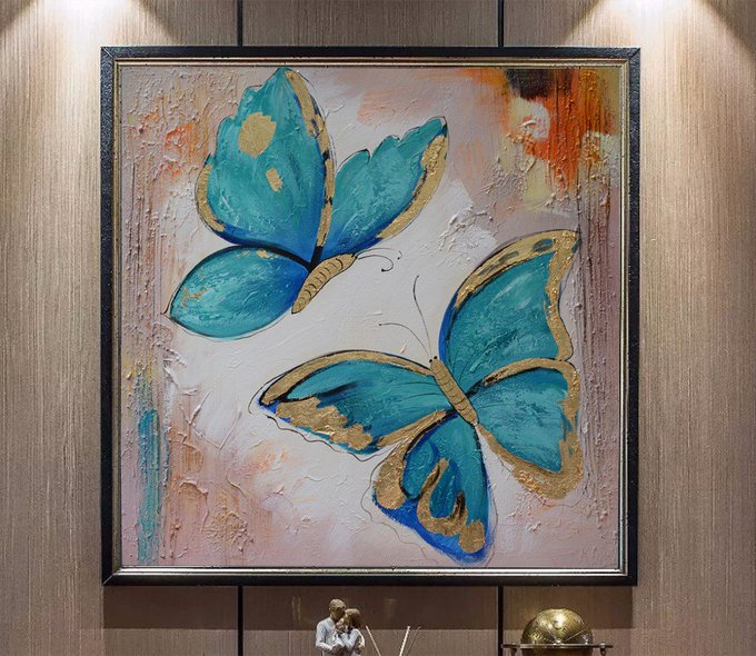 A framed textured painting of two teal blue butterflies with gold accents on a pink and orange background, hanging on a wooden wall above a small figurine and a golden globe ornament.