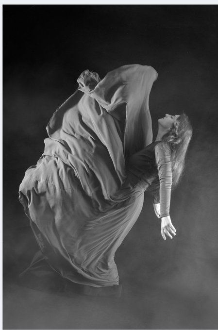 A dramatic black-and-white photograph of a dancer draped in flowing fabric, captured mid-movement with their body arched backward.