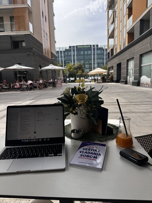 A laptop sits on an outdoor cafe table next to a book titled 'VESTINE VLADANE SOBOM' and a glass carafe of juice, with a 'Veletržní Banka' building visible in the background plaza.