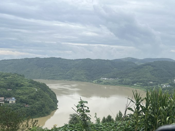 A scenic landscape photograph of a wide, muddy-colored river curving through lush green, forested mountains under a cloudy gray sky, likely taken from an elevated viewpoint.