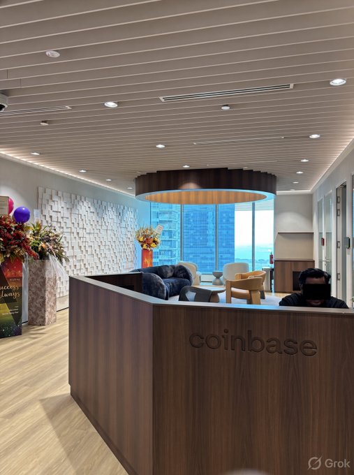 An office reception area features a wooden desk with the 'coinbase' logo, a person sitting behind it, and floral arrangements to the left; 'Grok' watermark in the corner.