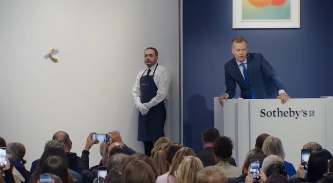 The image shows a group of people gathered around a man standing at a podium, possibly giving a speech or presentation. The attendees are using their phones to take pictures and record videos of the event. One person can be seen holding up their phone towards the camera. In the background, there is an art piece displayed on the wall that adds to the overall atmosphere of the scene.