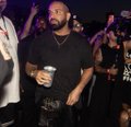The image shows the rapper Drake smiling and enjoying himself at what appears to be a lively nightclub or party event, holding a drink. He is surrounded by people, including someone in a white shirt visible on the left. The scene aligns with the post's caption suggesting that Drake is being gifted money by Stake, as he appears to be in a celebratory mood typical of his well-known appearances at Stake-sponsored events.