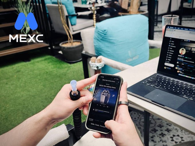 A person holds a smartphone displaying the MEXC crypto trading app while holding a hookah hose, with a laptop showing a trading interface on the table nearby; the MEXC logo is overlaid in the top-left corner.