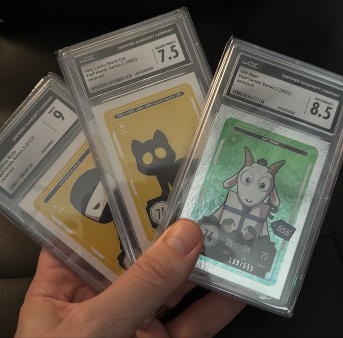 A hand holds three graded CGC trading cards in plastic slabs, including a 'Very Lucky Black Cat' card rated 7.5, a yellow card rated 9, and a 'Gift Goat' card rated 8.5.