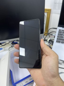 A hand holds a black smartphone with a dark screen, with a laptop and an HP monitor visible in the background on a desk.
