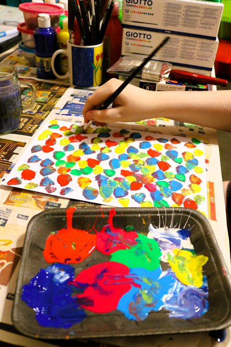 A child's hand stamps colorful paint onto paper, surrounded by a palette of vibrant red, blue, green, and yellow paints and GIOTTO brand supplies, capturing the "beautiful chaos" and creative energy referenced in the post about Team Cayden's art day.