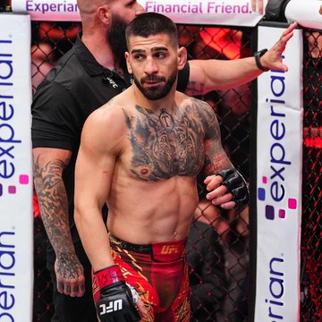 The image shows UFC fighter Ilia Topuria standing in an octagon with a championship belt, displaying a serious expression while wearing red and gold fight shorts. The image relates to the post as it depicts Topuria himself, who is the subject of speculation regarding his next opponent, with betting odds listing potential challengers including Paddy Pimblett, Justin Gaethje, Benoit Saint Denis, and Arman Tsarukyan.