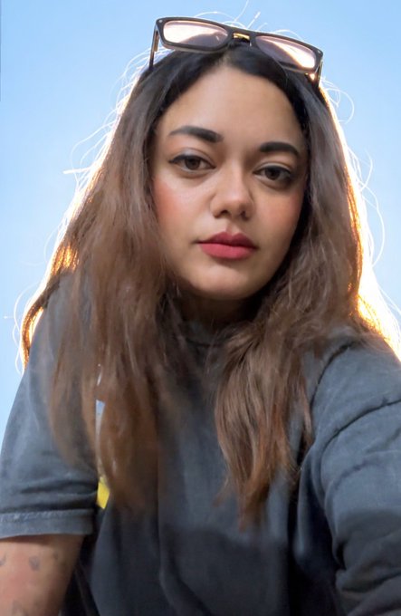 A selfie of a young woman with long brown hair, pink lipstick, and sunglasses perched on her head, wearing a dark gray t-shirt against a clear blue sky.