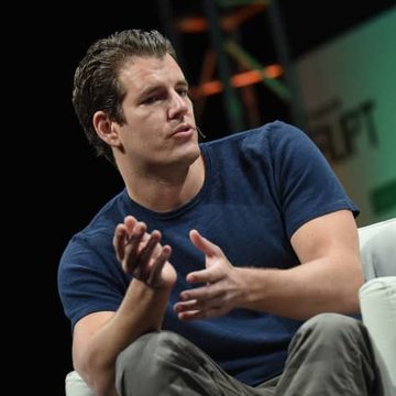 A man in a blue t-shirt sits on a white chair on a stage, gesturing with his hands as if speaking to an audience.
