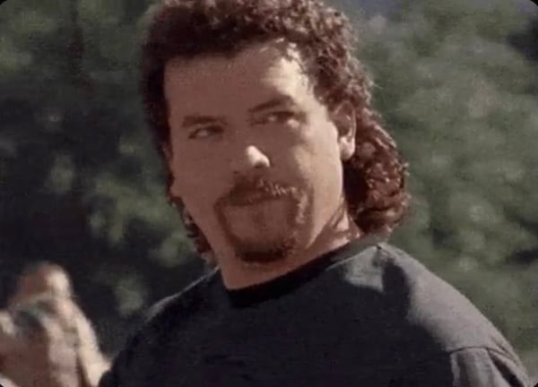 A close-up portrait of a man with curly dark hair and a goatee, wearing a dark shirt, with a serious expression and a blurred outdoor background.