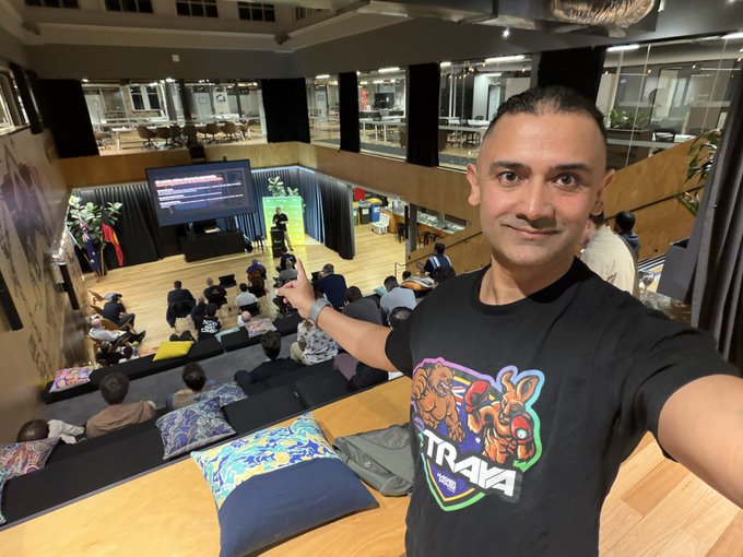 A man wearing a black 'STRAYA 100%' t-shirt takes a selfie while pointing toward an audience seated in a tiered auditorium, with a presentation screen visible on a stage below.