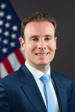 A professional headshot of a smiling man in a dark suit and light blue tie, with an American flag in the background.