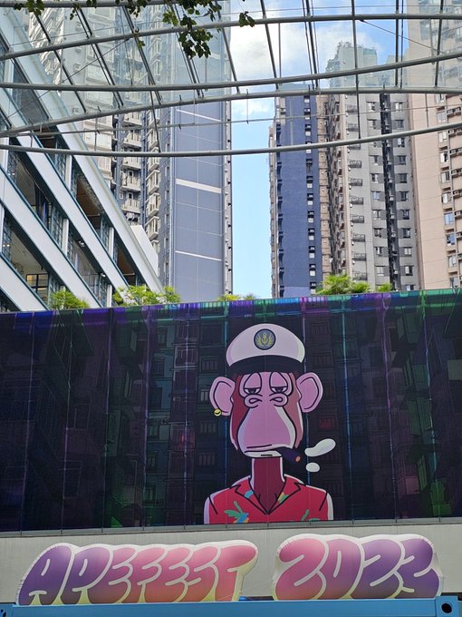 A large digital display showing an NFT-style cartoon monkey wearing a captain's hat and pink shirt, labeled 'APES3337 2023', set against a backdrop of high-rise buildings.