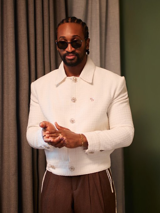 A stylish man with braided hair, a beard, and dark sunglasses stands confidently wearing an elegant white button-up jacket and brown pleated pants, hands clasped in front of him against a curtain backdrop. The caption "The Leader of the Trybe @trillionaire_V @encipherstudio #Chimsom #BBNaija" highlights his fashionable, leadership-style persona, matching the polished and composed look in the photo.