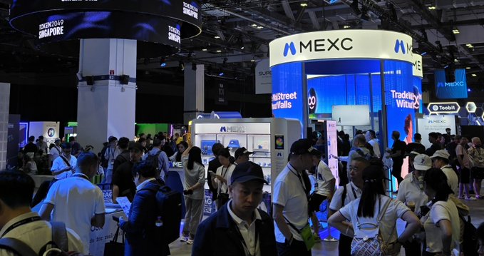 A crowded convention hall features a prominent illuminated "MEXC" booth displaying "MainStreet Wallets" and "Trade Your Winners," alongside "TOKEN2049 SINGAPORE" signage above the bustling attendees.