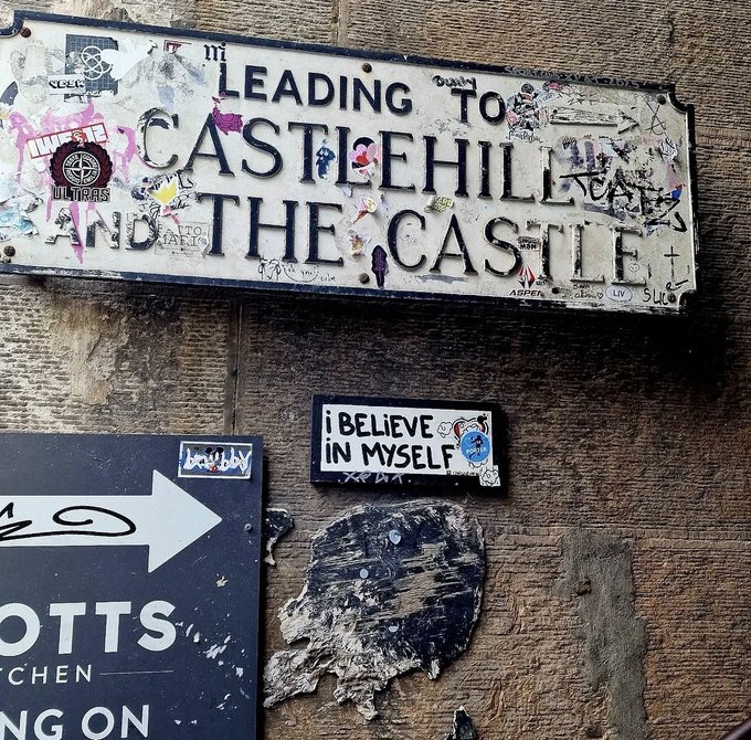 A photograph of signs mounted on a textured stone wall features a large, sticker-covered street sign reading 'LEADING TO CASTLEHILL AND THE CASTLE' positioned above a smaller sticker that says 'I BELIEVE IN MYSELF'.