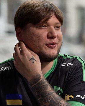 A close-up of a smiling man with a beard wearing a black and green esports jersey, featuring tattoos on his hand and neck.