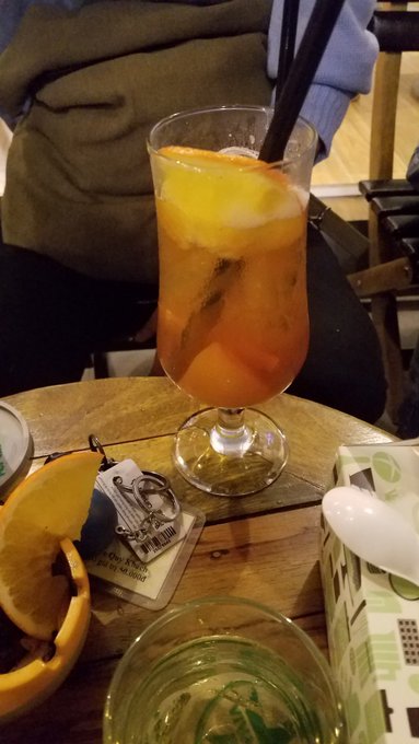 A tall glass of iced orange-colored beverage with an orange slice and black straw sits on a wooden table alongside a bottle opener with a keychain and an orange slice garnish.