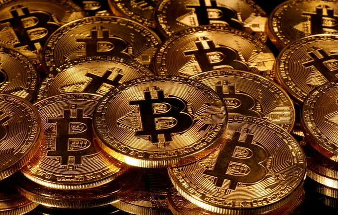 A pile of gold-colored Bitcoin tokens featuring the iconic "₿" symbol and partial inscriptions including "DIGITAL" and "DECENTRALIZED." The image illustrates the cryptocurrency's recent decline to around $77,100 amid broader risk-asset pressures from hot PPI data and climbing oil prices.