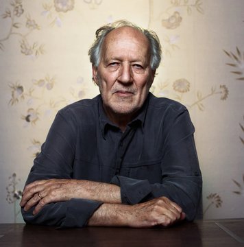 Portrait of an older man with gray hair wearing a dark shirt, sitting with crossed arms against a floral wallpaper background.