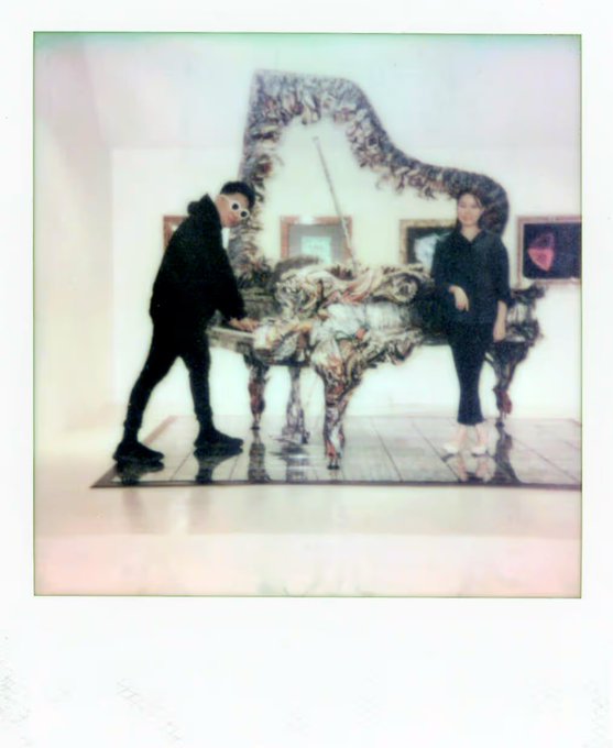 A Polaroid-style photograph showing two people standing on either side of a large sculptural art piece made of textured materials, displayed in what appears to be a gallery space.