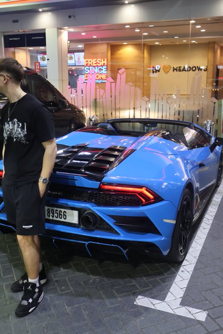 A bright blue Lamborghini convertible is parked on a city street near a building with signs reading 'FRESH SINCE 2000' and 'I ❤ MEADOWS', as a person in black shorts and shirt walks past.
