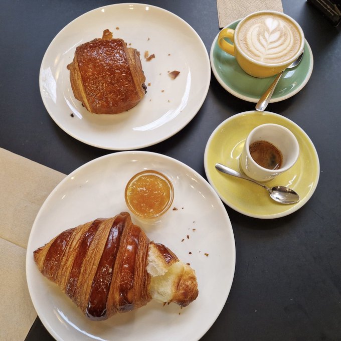 A breakfast spread featuring a chocolate-filled pastry and a croissant with a small jar of jam on white plates, accompanied by a yellow cup of latte and a small espresso.
