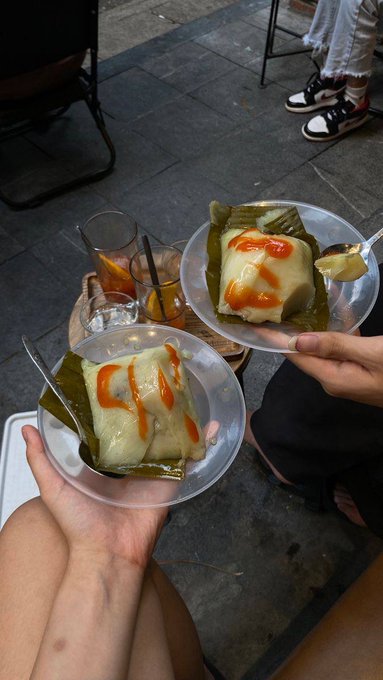 Two hands hold small bowls containing banana leaf-wrapped desserts topped with a red sauce, alongside glasses of iced tea, set on an outdoor stone surface.