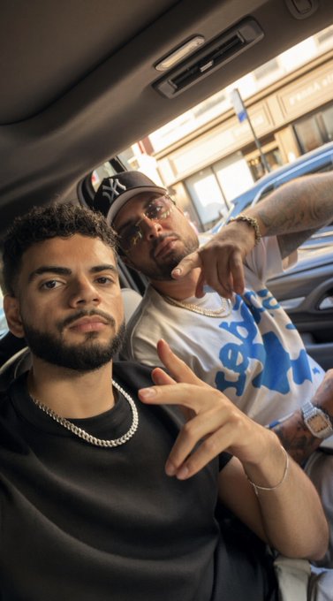 Two men sit inside a car; the man in the foreground wears a black shirt and silver chain, while the man in the back wears a New York Yankees cap, sunglasses, and a white and blue shirt.