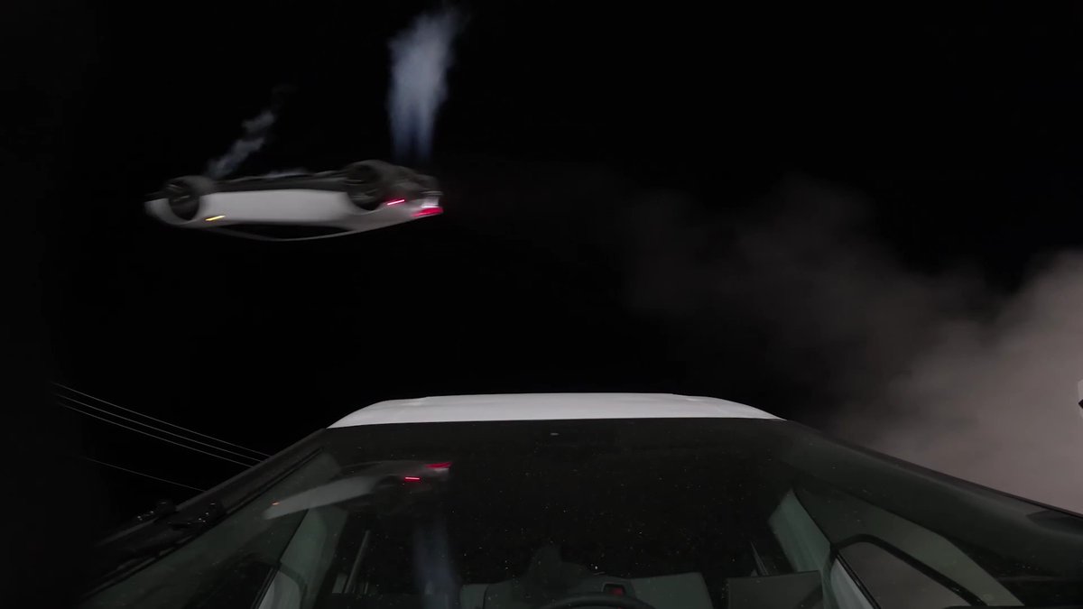 A nighttime scene showing a silver futuristic flying vehicle hovering in the dark sky above the roof of another vehicle, with smoke or mist visible around them.