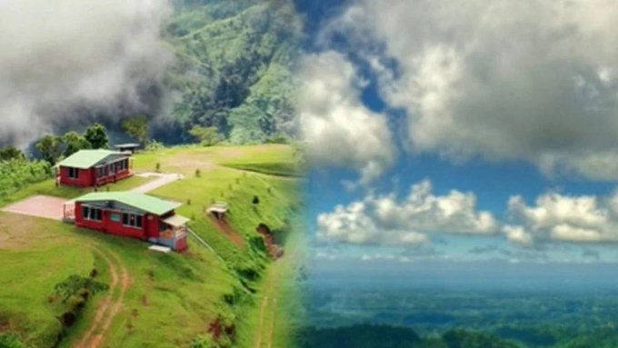 A split scenic view showing red cabins on a misty green hillside on the left and a bright blue sky with fluffy clouds over distant mountains on the right.